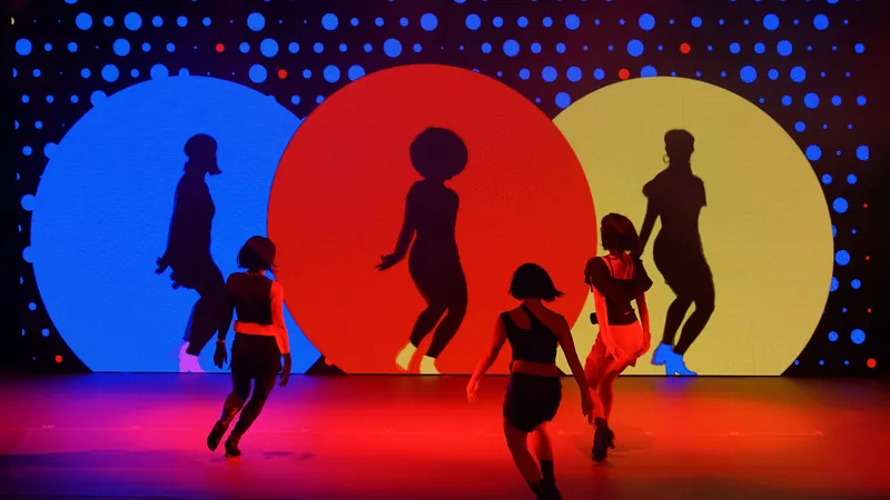Des danseurs en silhouette sur une scène, devant un mur d'écrans où se découpent trois grands cercles bleu, rouge et jaune.