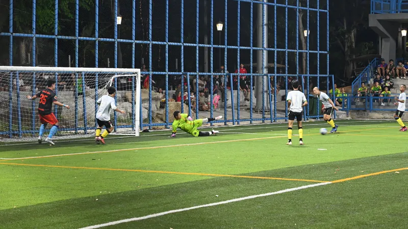 Match de football amateur en nocturne sur un terrain synthétique éclairé : le gardien plonge pendant qu'un joueur frappe au but.
