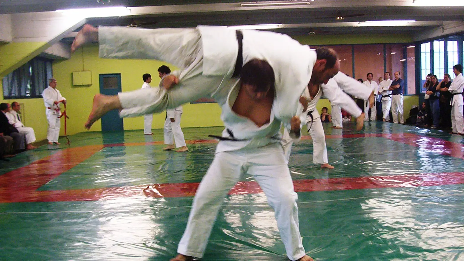 Dans un dojo, un judoka projette son partenaire par-dessus son épaule ; les autres élèves suivent la démonstration, assis au bord du tatami.